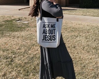 May include: A white tote bag with black straps and the text "Ask Me About Jesus" printed in black.