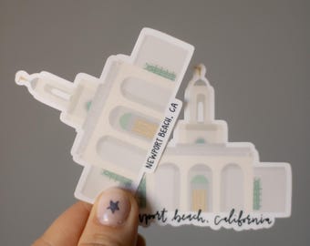 May include: A stack of white stickers featuring a stylized illustration of a building with arched windows and a domed roof. The stickers have the text "Newport Beach, CA" and "Newport Beach, California".