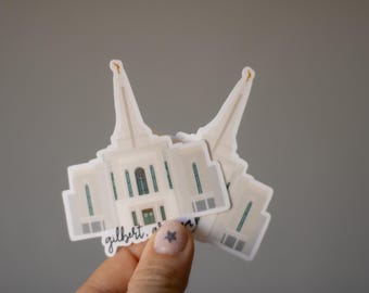 May include: Several white stickers featuring a detailed illustration of a building with two spires. The stickers have a light blue and green color scheme, with the text "gilbert, arizona" in a script font. The stickers are held against a gray background.