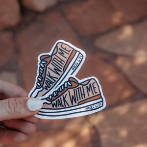 May include: Two stickers featuring a cartoon sneaker design in shades of orange and white. The text "WALK WITH ME" is prominently displayed, along with "MOSES G:34". The stickers are held by a person.