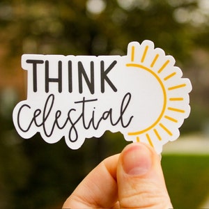 May include: A white sticker with the words "THINK celestial" in black and script font. A yellow sun graphic is on the right side. The sticker is held up by a person's hand against a blurred green background.