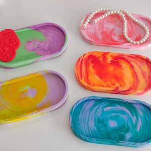 Op de afbeelding: Vijf ovale harsbakken met een marmerpatroon. De bakken zijn in verschillende kleuren, waaronder groen, paars, geel, oranje en blauw. De bakken zijn perfect om sieraden, sleutels of andere kleine voorwerpen in te bewaren.