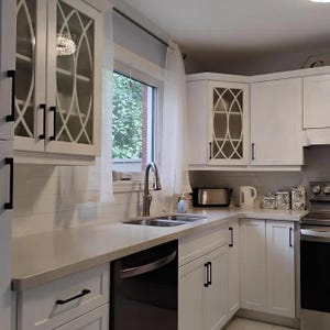 May include: A kitchen featuring white cabinets with black hardware and glass-paneled doors displaying an oval lattice design. The space includes a stainless steel faucet, a double-basin sink, a black dishwasher, and a white subway tile backsplash.