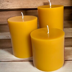 May include: Three yellow beeswax candles of varying sizes on a wooden surface. The candles are unlit and have white wicks.