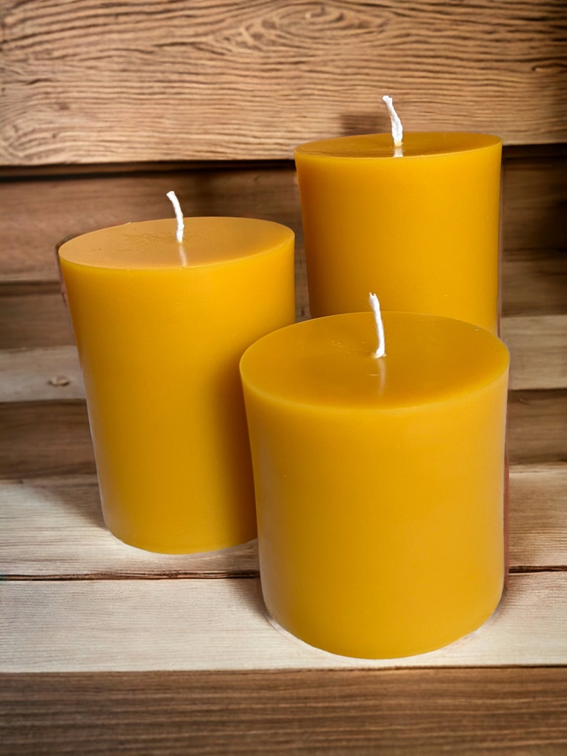 May include: Three yellow beeswax candles of varying sizes on a wooden surface. The candles are unlit and have white wicks.