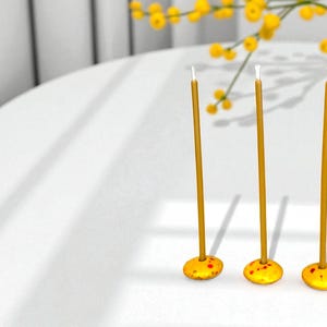May include: Three tall, slender, yellow candles with white wicks stand on a white table. Each candle is set in a small, round, yellow base with red speckles. A vase of yellow flowers is in the background.