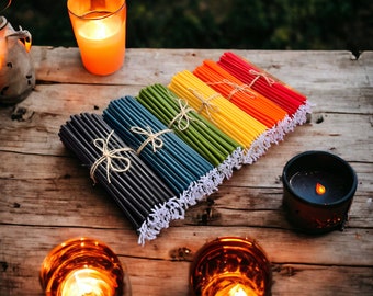 Op de afbeelding: Een bundel kleurrijke bijenwaskaarsen, gebonden met touw, gerangschikt in een regenboogpatroon op een houten oppervlak. De kaarsen zijn in verschillende tinten rood, oranje, geel, groen, blauw en zwart.