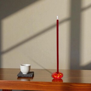 May include: A minimalist still life featuring a red incense stick with a decorative base, a white teacup, and a small square coaster. The objects are arranged on a wooden table, with shadows cast on the wall.