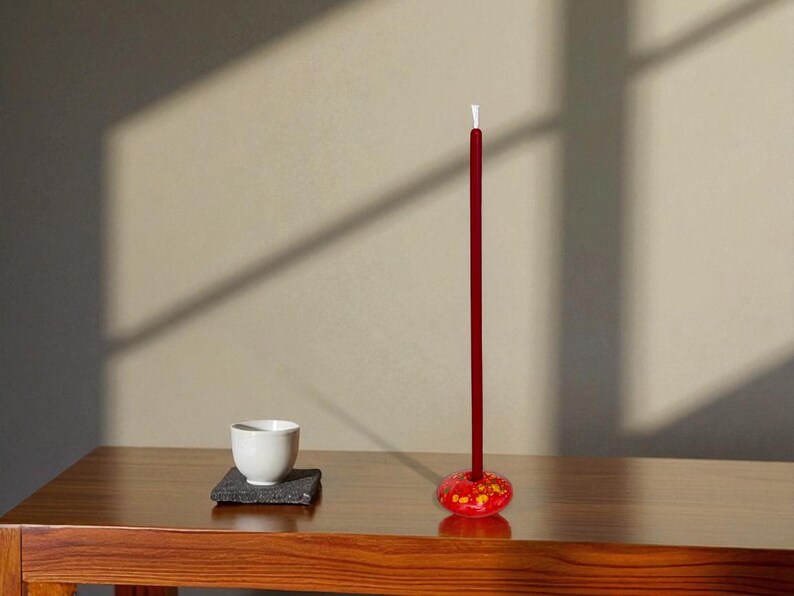 May include: A minimalist still life featuring a red incense stick with a decorative base, a white teacup, and a small square coaster. The objects are arranged on a wooden table, with shadows cast on the wall.