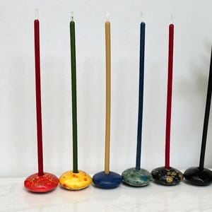 May include: A collection of six tall, slender candles in various colors. Each candle is mounted on a decorative, rounded base. The candles are red, green, tan, blue, and black. The bases are painted in complementary colors and patterns.
