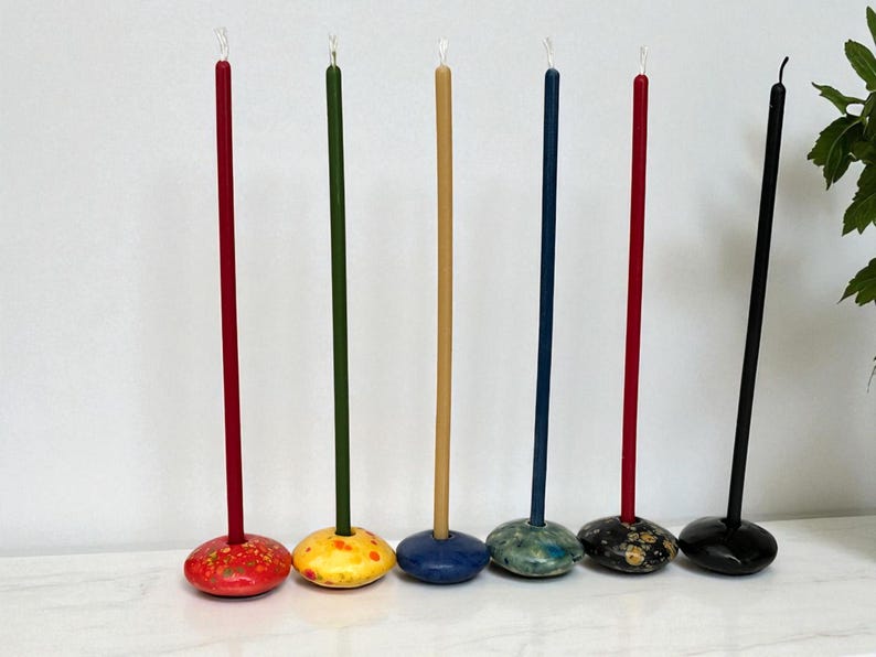 May include: A collection of six tall, slender candles in various colors. Each candle is mounted on a decorative, rounded base. The candles are red, green, tan, blue, and black. The bases are painted in complementary colors and patterns.