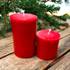 May include: Two red pillar candles, one larger than the other, on a wooden surface. The candles are unlit and have a white wick.