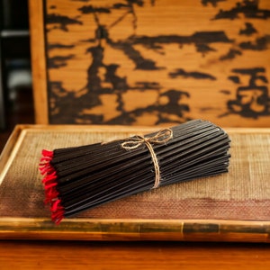 May include: A bundle of black incense sticks tied together with twine and red string. The incense is sitting on a wooden tray with a woven pattern.