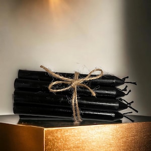 May include: A bundle of black taper candles tied together with twine. The candles are arranged in a stack and are ready to be used for rituals or spell work.