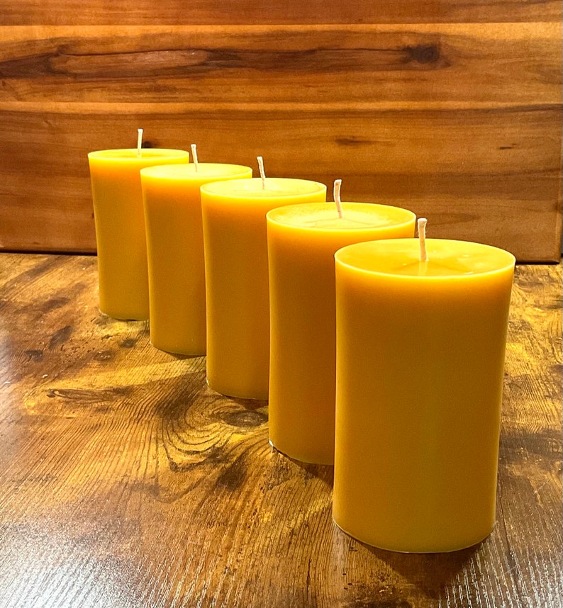 May include: Four yellow beeswax candles arranged in a row on a wooden surface. The candles are all the same size and shape, and they have a natural, rustic look.