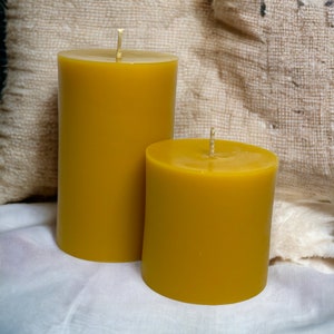 May include: Two yellow beeswax candles, one larger than the other, on a white fabric surface. The candles are unlit and have wicks.