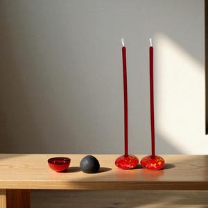 May include: A minimalist still life featuring two tall, red candles with white tips, set in decorative holders with red and yellow floral patterns. A small red bowl and a dark gray stone sit nearby on a wooden surface.