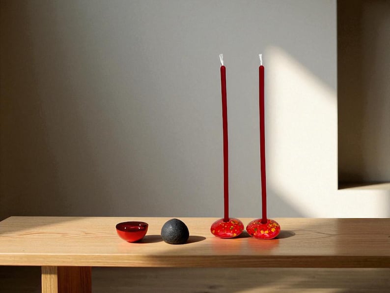 May include: A minimalist still life featuring two tall, red candles with white tips, set in decorative holders with red and yellow floral patterns. A small red bowl and a dark gray stone sit nearby on a wooden surface.
