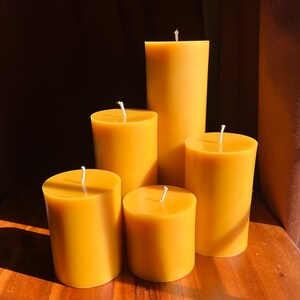 May include: Four yellow beeswax candles of varying sizes, arranged on a wooden surface. The candles are all cylindrical in shape and have a natural, unbleached color.