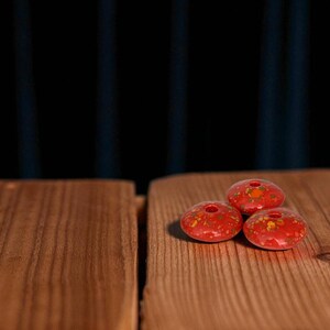 May include: Three red, ceramic beads with a speckled, yellow and orange pattern. Each bead has a central hole. The beads are displayed on a wooden surface with a dark blue background.
