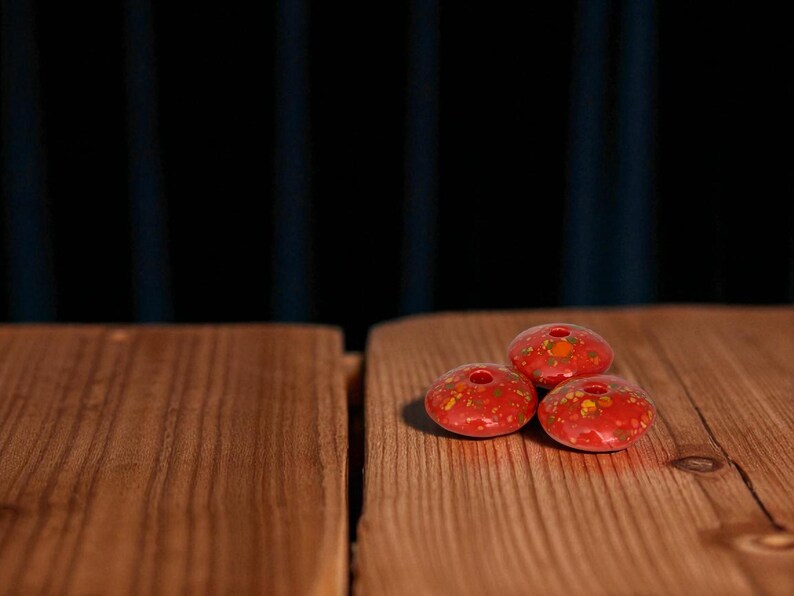 May include: Three red, ceramic beads with a speckled, yellow and orange pattern. Each bead has a central hole. The beads are displayed on a wooden surface with a dark blue background.
