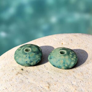 May include: Two turquoise ceramic beads with a speckled pattern and a central hole, resting on a light-colored stone. The beads have a slightly irregular, organic shape, with a glossy finish.