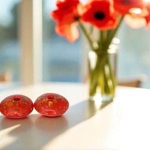 May include: Two red, decorative glass objects with a hole in the center, speckled with yellow and orange, sit on a white table. A vase of red poppies is in the background. The scene is lit by sunlight.