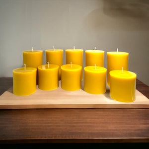 May include: Ten yellow beeswax candles arranged in a row on a wooden surface. The candles are all the same size and shape, and they are all lit.