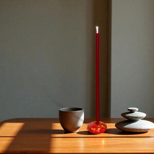 May include: A minimalist still life featuring a red incense stick with a decorative base, a dark ceramic cup, and a stack of smooth stones. The objects are arranged on a wooden surface, with sunlight casting shadows.