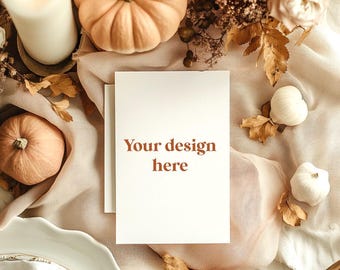 May include: A white rectangular greeting card with the text Your design here in brown font, surrounded by light beige fabric, small white and orange pumpkins, a tall white pillar candle, and dried autumn leaves on a white plate.