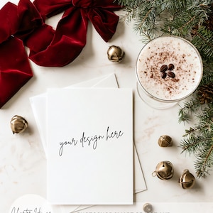 May include: A white rectangular greeting card mockup with the text your design here in black script font. The card is surrounded by a large red velvet bow, a glass of creamy coffee with beans, several small gold jingle bells, and pine branches.