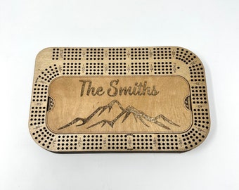 May include: A wooden personalized cribbage board with a mountain range design and the text "The Smiths". The board has a grid of holes for scoring and a space for the pegs.