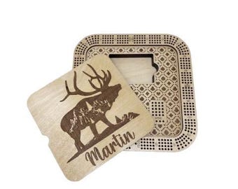 May include: A wooden cribbage board with a decorative elk design. The board has a square shape with a patterned border and a sliding cover. The elk design is on a separate wooden piece with the name "Martin" engraved.