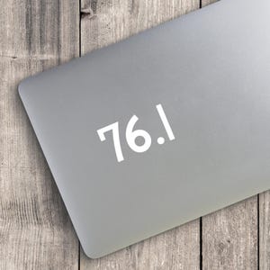 May include: A matte gray laptop lid featuring a white vinyl decal of the number 76.1 in a clean, sans-serif font. The decal is centered on the smooth, metallic surface of the laptop, which is positioned at an angle against a wooden background.