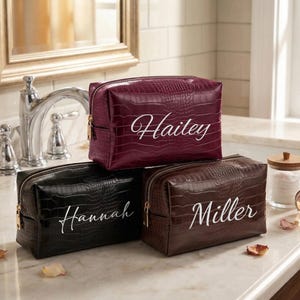 May include: Three personalized cosmetic bags in burgundy, black, and brown, each with a reptile skin texture and a gold zipper. The bags are labeled with the names "Hailey", "Hannah", and "Miller" in white script.