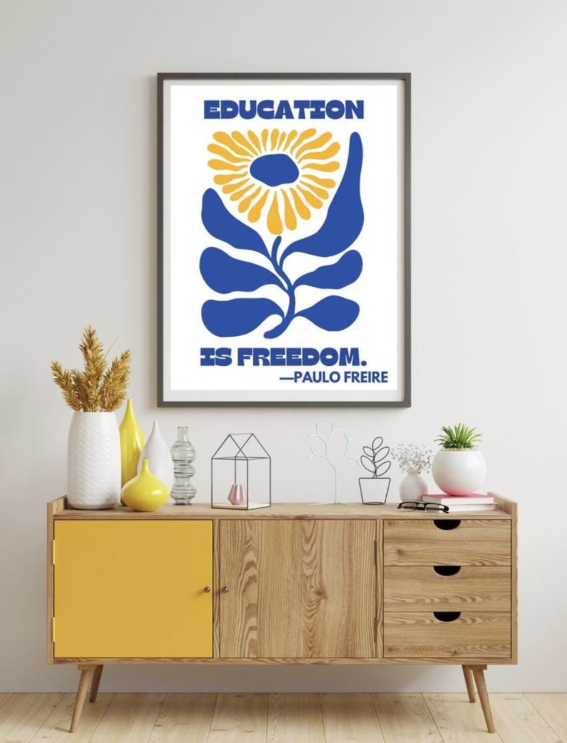 Pu&ograve; includere: Un poster incorniciato con un design floreale blu e giallo. Il poster recita "EDUCATION IS FREEDOM. -PAULO FREIRE".