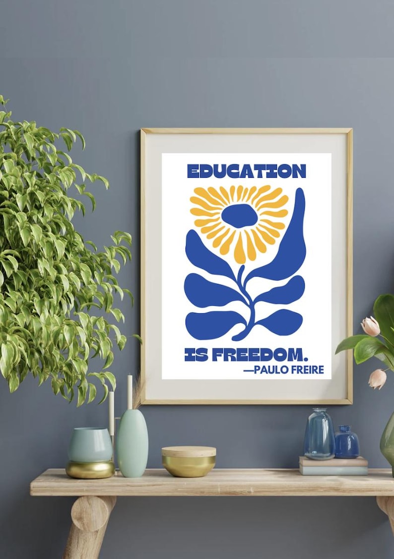 Pu&ograve; includere: Una stampa incorniciata con uno sfondo bianco e un fiore stilizzato blu con un centro giallo. Il testo "EDUCATION IS FREEDOM." &egrave; stampato sotto il fiore, con la citazione "PAULO FREIRE" sotto.