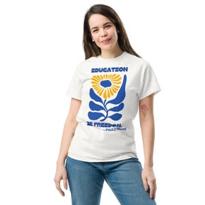 May include: White t-shirt featuring a blue and yellow floral design with the words "EDUCATION IS FREEDOM" and the name "PAULO FREIRE" in blue text. The shirt is made of a soft material and is being worn.