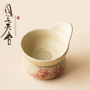 May include: A beige ceramic tea strainer with a handle, set atop a matching base. The strainer has a perforated bottom and a slightly textured surface. The base features a reddish-brown floral design. Chinese characters are visible on the left.
