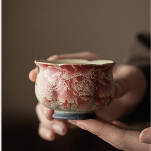 May include: A small, ceramic teacup with a floral design. The cup features a red and white peony pattern on a cream-colored background. The rim is scalloped, and the base is a dark blue. The cup is held in a person's hands.