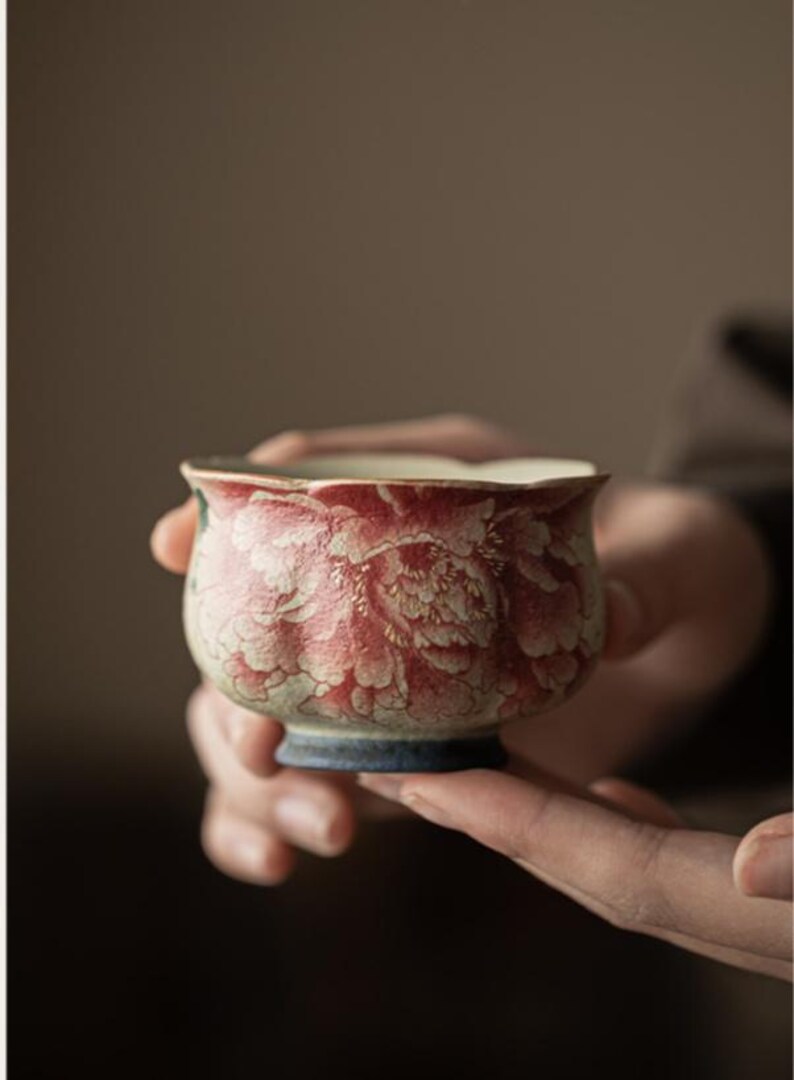 May include: A small, ceramic teacup with a floral design. The cup features a red and white peony pattern on a cream-colored background. The rim is scalloped, and the base is a dark blue. The cup is held in a person's hands.
