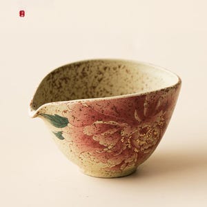 May include: A ceramic tea pitcher with a speckled beige exterior and a pink floral design. The pitcher features a spout and a wide opening. The floral design includes pink petals and green leaves, with gold accents.