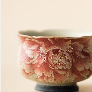 May include: A ceramic bowl with a floral design in shades of red and white, accented with gold details. The bowl has a rounded shape with a dark blue base and a light-colored interior. The floral pattern covers the exterior.