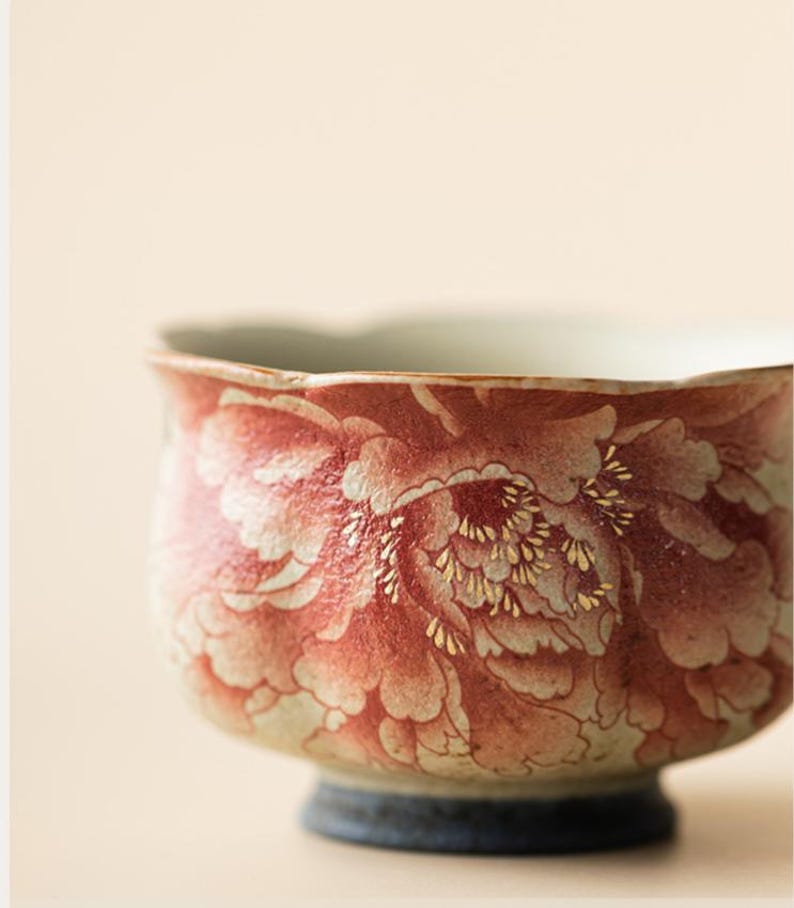 May include: A ceramic bowl with a floral design in shades of red and white, accented with gold details. The bowl has a rounded shape with a dark blue base and a light-colored interior. The floral pattern covers the exterior.