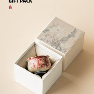 May include: A white gift box with a floral design on the lid, open to reveal a small, decorative ceramic cup with a red floral pattern. The text "GIFT PACK" is at the top.