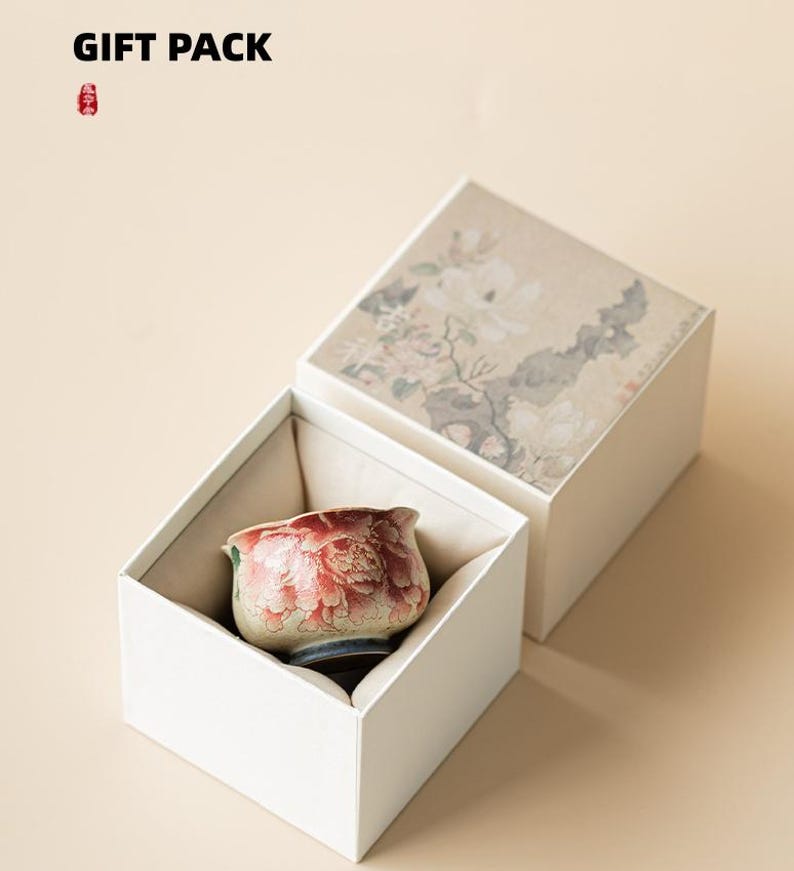 May include: A white gift box with a floral design on the lid, open to reveal a small, decorative ceramic cup with a red floral pattern. The text "GIFT PACK" is at the top.