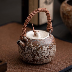 Könnte beinhalten: Eine braune Teekanne aus Keramik mit einem weißen Blumenmuster und einem geflochtenen Griff. Die Teekanne hat einen weißen Deckel mit einem kleinen Knopf.