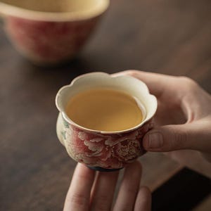 May include: A small, floral-patterned teacup filled with tea, held in a person's hands. The cup features a red and white floral design with a scalloped rim. The tea inside is a light amber color. Another teacup is slightly out of focus in the background.