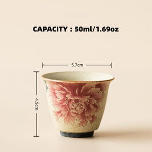 May include: A ceramic teacup with a floral peony design in shades of red and pink. The cup is 4.5 cm tall and 5.7 cm wide, with a capacity of 1.69 oz. The text "CAPACITY: 50ml/1.69oz" is displayed above the cup.