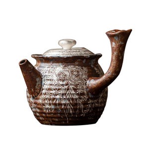 Könnte beinhalten: Eine braune Teekanne aus Keramik mit einem silbernen Deckel und einem floralen Design. Die Teekanne hat einen Ausguss und einen Griff.